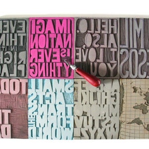 May include: A set of nine woodblock printing stamps with various phrases and words, including "Everything is in the right place", "Imagination is everything", "Hello, so love you", "Be so 2020", "Small acts for the world", "Today is the day", and a world map. A red carving tool is placed on top of the stamps.