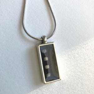 May include: Silver-toned necklace with a rectangular pendant. The pendant has a dark, possibly black, background with three small, square, silver-colored accents. The chain is a thin, snake chain style.