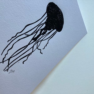 May include: A black jellyfish linocut print on white paper. The jellyfish is depicted with a rounded bell and long, flowing tentacles. The print is signed in the bottom left corner. The artwork is a minimalist representation of marine life.