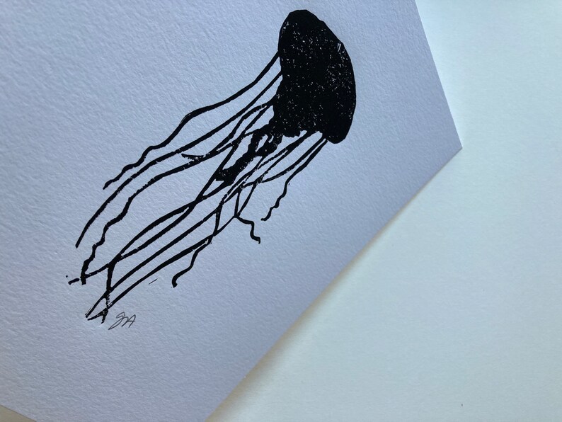 May include: A black jellyfish linocut print on white paper. The jellyfish is depicted with a rounded bell and long, flowing tentacles. The print is signed in the bottom left corner. The artwork is a minimalist representation of marine life.