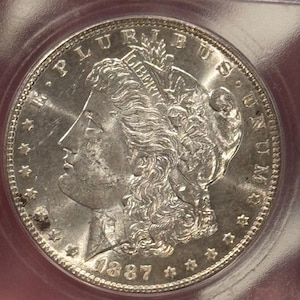 May include: A silver Morgan dollar coin with the year 1887 inscribed on the front. The coin features a portrait of Lady Liberty with flowing hair and a crown.