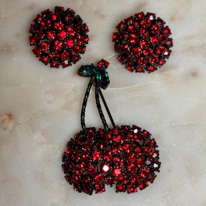 Peut inclure: Un ensemble de bijoux vintage sur le thème de la cerise, comprenant une broche et des boucles d'oreilles assorties. La broche et les boucles d'oreilles sont incrustées de strass rouges vifs, avec un accent de strass vert sur la broche. Les tiges sont noires.