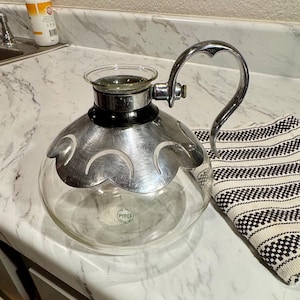 Può includere: Una caffettiera Pyrex vintage con corpo in vetro trasparente e una fascia decorativa in metallo argentato. La caffettiera ha un manico a forma di cuore. C'è un panno a righe bianche e nere accanto. Il logo Pyrex è visibile.