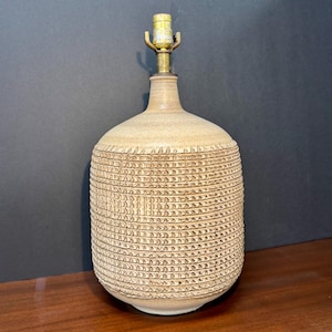 May include: A beige ceramic lamp base with a cylindrical body featuring a repeating horizontal textured pattern of small indentations. The lamp has a narrow neck and a brass-colored metal socket fixture at the top, centered on the rounded shoulder.