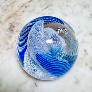 Puede incluir: Un pisapapeles esférico de cristal transparente con un diseño azul y blanco. El pisapapeles presenta un patrón arremolinado de rayas azules y blancas y un área esmerilada texturizada. Se pueden ver pequeñas burbujas de aire dentro del cristal.