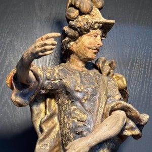May include: A vintage, hand-carved wooden statue of a figure in antique attire. The statue features a hat with decorative elements, a mustache, and a detailed facial expression. The color palette includes gold, brown, and red accents, with visible signs of age and wear.