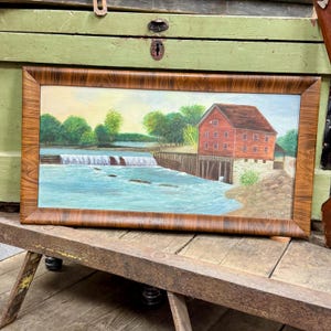 May include: A framed landscape painting depicts a red mill building beside a waterfall. The water is blue, and the sky is a mix of yellow and blue. The painting is in a brown wooden frame.