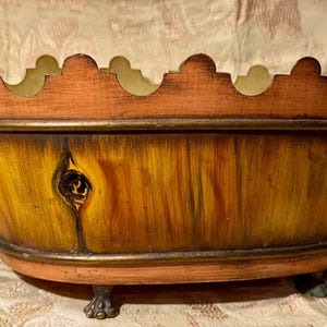 Puede incluir: Un macetero ovalado con diseño de veta de madera, con un borde de color cobre y asas decorativas de color dorado. El macetero tiene tres patas y un borde festoneado.
