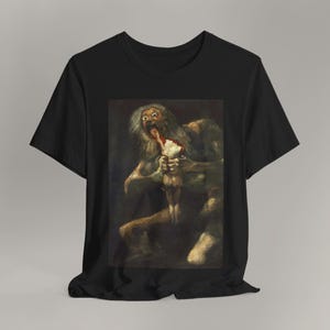 Könnte beinhalten: Ein schwarzes Kurzarm-T-Shirt mit einem rechteckigen Grafikdruck von Francisco Goyas Gemälde Saturn verschlingt seinen Sohn. Das Kunstwerk zeigt eine große, wild blickende Figur mit grauem Haar, die eine kleinere Figur vor dunklem Hintergrund verzehrt.