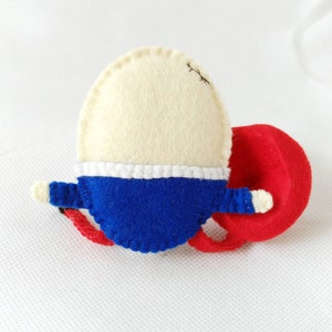 Puede incluir: Un broche de fieltro hecho a mano con forma de velero. La vela es de color crema, el casco es azul con una franja blanca y la boya es roja. El broche tiene peque&ntilde;os detalles blancos y costuras negras.