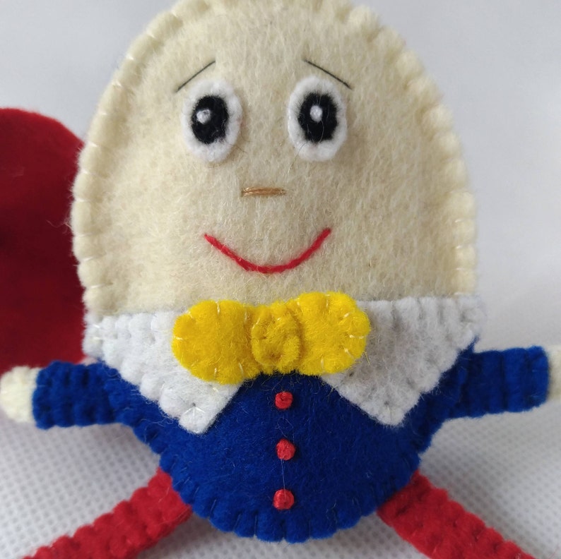 Puede incluir: Mu&ntilde;eco de fieltro hecho a mano de Humpty Dumpty. El mu&ntilde;eco tiene una cabeza en forma de huevo de color crema con ojos negros, una sonrisa roja y una pajarita amarilla. Lleva una chaqueta azul con botones rojos, un cuello blanco y piernas rojas.