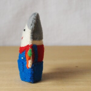 May include: A handmade felt doll with a white face, gray hat, and red shirt. The doll wears blue overalls and has a small, colorful accessory. The doll is standing on a wooden surface.