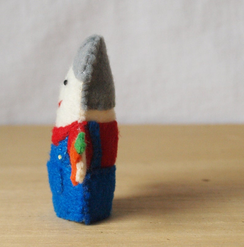 May include: A handmade felt doll with a white face, gray hat, and red shirt. The doll wears blue overalls and has a small, colorful accessory. The doll is standing on a wooden surface.