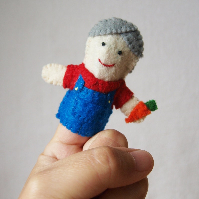 May include: A handmade felt finger puppet of a smiling farmer. The puppet wears a red shirt, blue overalls, and a gray cap. It holds a small orange carrot with a green top. The puppet is designed to fit on a finger.
