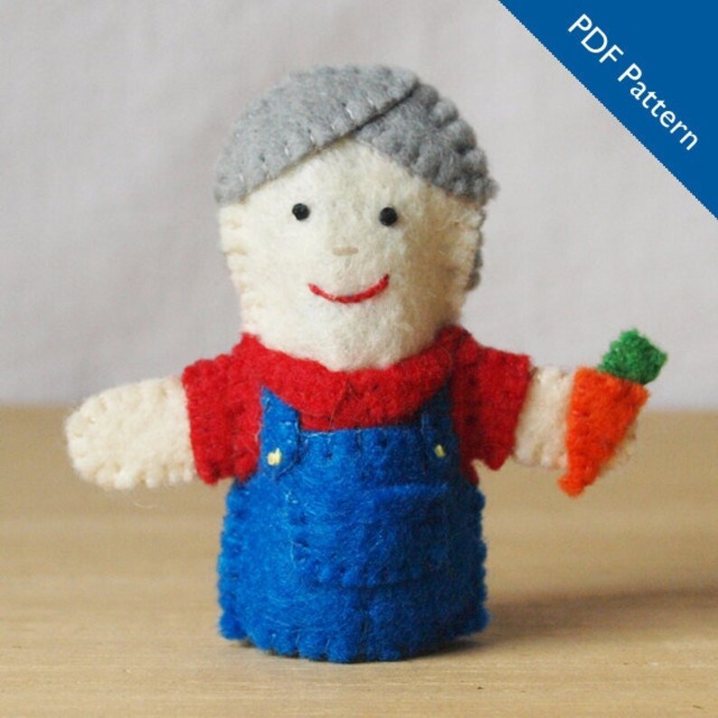 May include: A handmade felt puppet of a person wearing a red shirt and blue overalls, holding a felt carrot. The puppet has gray hair and a smiling face. The words "PDF Pattern" are in the upper right corner.