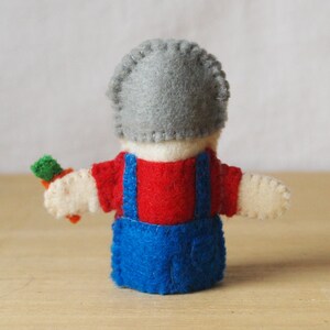 May include: A handmade felt doll, viewed from the back, wearing a gray hat, red shirt, and blue overalls. The doll is holding a small carrot. The doll's arms are outstretched, and the stitching is visible.