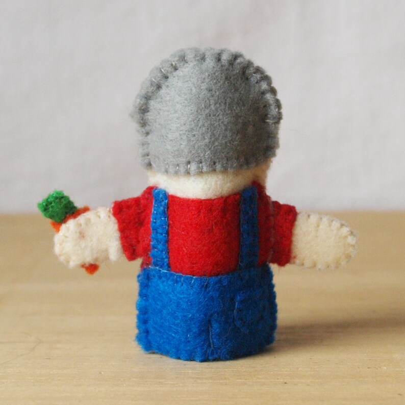 May include: A handmade felt doll, viewed from the back, wearing a gray hat, red shirt, and blue overalls. The doll is holding a small carrot. The doll's arms are outstretched, and the stitching is visible.