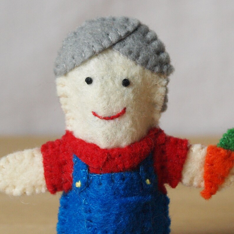 May include: A handmade felt doll depicting a farmer. The doll has a cream-colored face, gray hair, a red shirt, blue overalls, and a smiling face. The doll is holding a carrot.