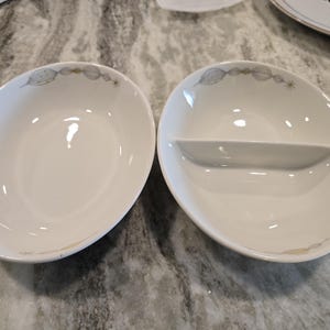 May include: Two white oval serving bowls with a decorative leaf and dot pattern around the rim. One bowl is divided into two sections. The bowls are on a gray and white surface.