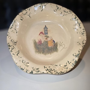May include: Cream-colored ceramic bowl with a scalloped edge, speckled with green. The interior features a hand-painted scene of birdhouses and foliage. A signature is visible on the right side of the bowl.
