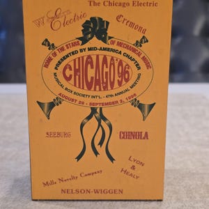 May include: A vintage matchbox with a yellow background and brown trim. The box features text and graphics for the "Chicago 96" Musical Box Society Int'l. meeting, including dates and company names. The design includes ribbons and musical instrument illustrations.