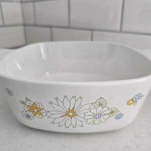 May include: White square baking dish with a floral daisy design in yellow, green, and blue. The dish has a simple, clean design, perfect for baking and serving. The dish is made of a durable material.