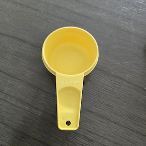 May include: A yellow plastic measuring cup with a handle. The cup is labeled "1/3 CUP". The handle has a hole at the end. The cup is sitting on a dark gray wooden surface.