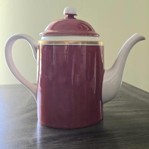 May include: A burgundy and white ceramic teapot with a gold trim. The teapot has a curved handle and spout, and a lid with a white knob. The teapot is on a dark surface.