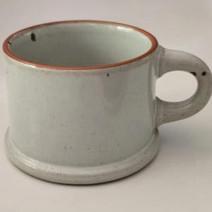 May include: A light gray ceramic mug with a speckled finish and a thick, rounded handle. The mug features a distinct, raised base and a thin, reddish-brown rim around the top edge. The overall surface has a smooth, matte texture with small dark spots.