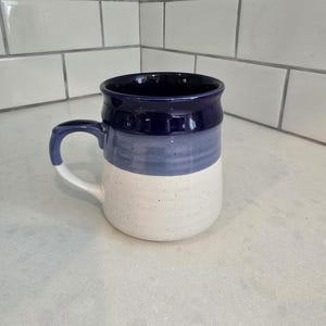 May include: A handmade ceramic mug with a white speckled base, a light blue middle section, and a dark blue rim and handle. The mug is designed with a curved handle for easy gripping and is suitable for hot or cold beverages.