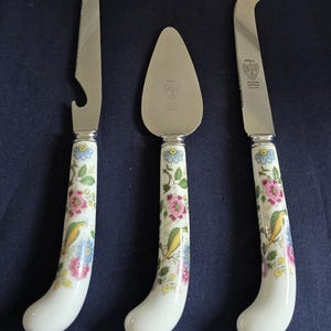May include: A set of three stainless steel serving utensils with white handles. The handles are decorated with a floral and bird pattern in pink, blue, and yellow. The set includes a cheese knife, a cake server, and a cheese knife with a serrated edge.