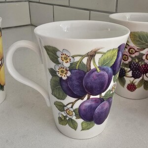 May include: White ceramic mug with a curved handle, decorated with a plum branch design. The plums are a deep purple, complemented by white blossoms and green leaves. Another mug with a similar design is partially visible.