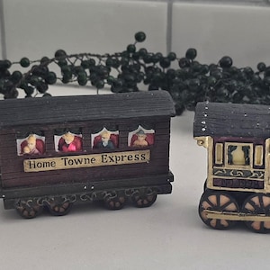 May include: Three miniature train cars. The train cars are painted in shades of brown, tan, and black. The middle car has the words "Home Towne Express" on the side. The train cars are decorated with small figures.