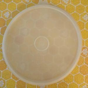 May include: A round, translucent, off-white plastic lid featuring a subtle, embossed honeycomb pattern across its surface. The lid includes a small, centered circular indentation and a single tab at the top edge for easy removal from a container.