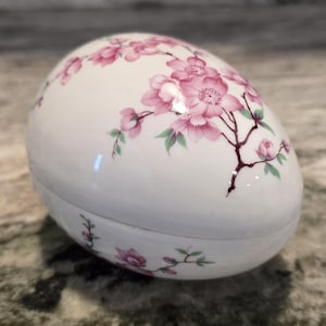 May include: White ceramic egg-shaped trinket box with a lid. The box is decorated with pink cherry blossom branches and green leaves. The box is likely used for storing small jewelry or keepsakes.