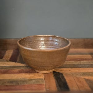 May include: A handmade, brown ceramic bowl with a textured surface. The bowl has a rounded shape and a slightly rough, unglazed base. The interior of the bowl has a glossy finish, reflecting light. This bowl is suitable for serving food.