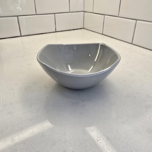 May include: A gray, square-shaped ceramic bowl with gently curved sides. The bowl is set against a white countertop and a backdrop of white subway tiles. The bowl's interior reflects light, highlighting its smooth surface.