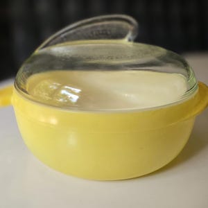 Könnte beinhalten: Eine gelbe, runde Vintage-Küchenschüssel mit einem Deckel aus klarem Glas. Der Deckel hat einen Griff. Die Schüssel hat zwei kleine Griffe auf beiden Seiten. Die Schüssel steht auf einer weißen Oberfläche.