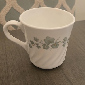 May include: White ceramic mug with a curved handle and a decorative vine and leaf pattern in shades of green and gold. The mug has a spiral design on the exterior.