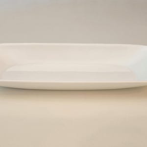 May include: A rectangular white ceramic serving platter with rounded corners and slightly raised edges. The dish features a smooth, glossy finish and a shallow, flat base, presenting a clean and simple design for serving various food items.