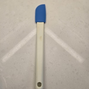 May include: A blue and white silicone spatula. The spatula has a blue, rounded head and a white handle with a hole at the end. The spatula is designed for cooking and baking.