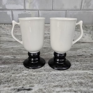 May include: Two white ceramic mugs with black pedestal bases. The mugs have a classic design with a curved handle. The bases are a glossy black, and the mugs are set against a neutral background.