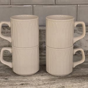 May include: Four stacked off-white ceramic mugs with a vertical ribbed pattern. Each mug has a curved handle and a cylindrical shape. The mugs are arranged in two stacks of two, with the handles facing outward.