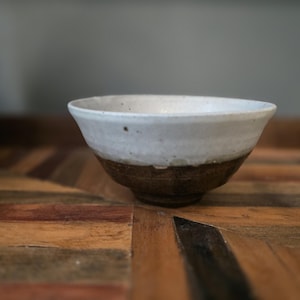 May include: A handcrafted ceramic bowl, white at the top and dark brown at the base. The bowl has a wide, slightly flared rim and sits on a wooden surface. The bowl's exterior has a textured, rustic appearance.
