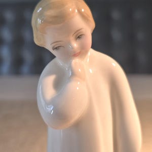 May include: A porcelain figurine of a young person, with a serene expression. The figure is dressed in a white robe, with a hand gently touching the chin. The hair is a light blonde color, and the skin has a delicate, painted finish. The figurine is a decorative item.