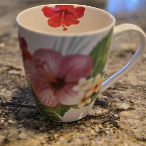 Pode incluir: Caneca de cerâmica branca com um design floral com flores de hibisco rosa, vermelhas e amarelas e folhas verdes.