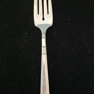 May include: A silver-toned metal fork featuring four tines and a slender handle. The handle is decorated with a geometric pattern consisting of small squares and a repeating diamond motif near the neck, ending in a flat, squared-off base.