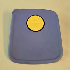 May include: A square, periwinkle blue Tupperware storage container lid featuring a circular, bright yellow vent button in the center. The lid has rounded corners and a small, raised tab on one corner for easy removal from the container base.