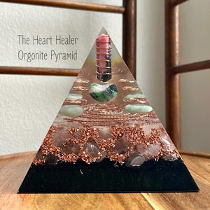 May include: Orgonite pyramid with a black base, copper wire, and embedded crystals. The pyramid features a pink and green stone, a green heart-shaped stone, and the text "The Heart Healer Orgonite Pyramid".
