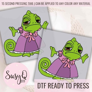 May include: A green chameleon wearing a purple dress with a pink bow. The chameleon is in a cartoon style and has a sad expression. The text "DTF READY TO PRESS" is below the chameleon.